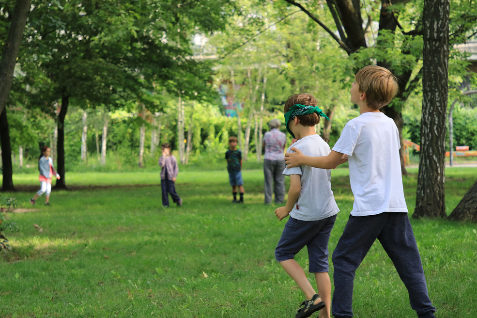 Ein Junge führt einen anderen Jungen, dessen Augen verbunden sind durch ein Feld mit Bäumen.